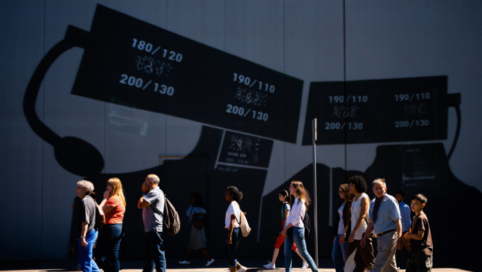 Una folla di persone cammina sotto l'ombra di un simbolo di ipertensione, a rappresentare i nuovi malati a causa delle nuove linee guida.